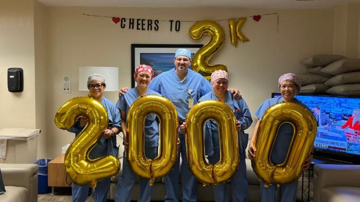 Five nurses and doctors in blue scrubs holding number balloons to show the number two thousand. They are standing in a dimly lit lounge room with beige couches. 
