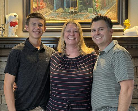 A family of three - two parents and a son - stand in front of a fireplace with two snoopy toys and a painting hanging over it. 