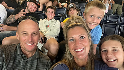 A middle aged woman sits with her husband and two children in bleachers while watching a baseball game.