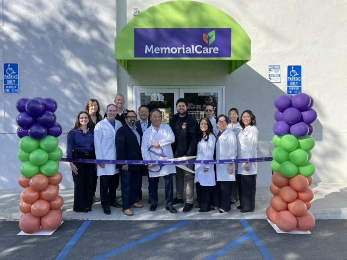 A group of doctors, city officials, and heath care executives standing between two balloon pillars with purple, green and orange balloons, with the center doctor holding giant scissors to cut a purple ribbon with the MemorialCare logo on it. 
