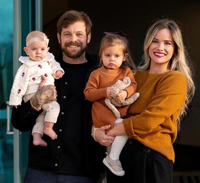 A man and woman hold their two children, the woman is wearing an orange sweater and holding a small child wearing a matching sweater, while the man wears a black sweater while holding a child in a white sweater and wihte bow. 