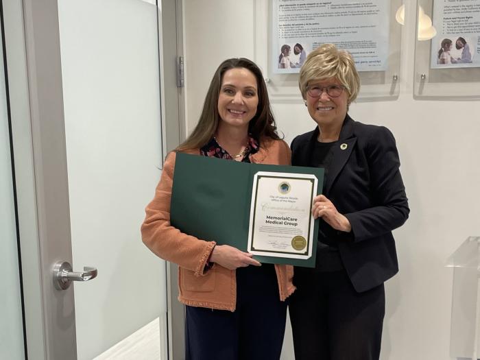 Two women (one in a brown jacket and black pants, and one in a black blazer, black shirt and black pants) stand next to a door in a medical clinic holding a forest green certificate of commendation to MemorialCare's commitment to the Laguna Woods community.  
