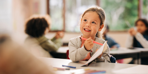 Six year old with pen and paper sitting and smiling at school desk.