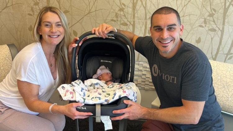 Kirsten Ernst and her husband sit on comfy chairs while holding their newborn in a car seat. 