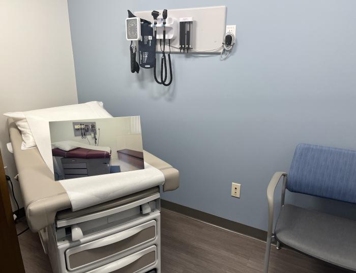 A newly remodeled medical examination room with medical bed, chair, blood pressure monitor and eye/throat instruments with a picture of the old design of the room sitting on the bed. 