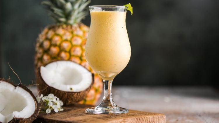 A tropical colada in a glass cup placed on top of a wooden board surrounded by coconuts and pineapples.